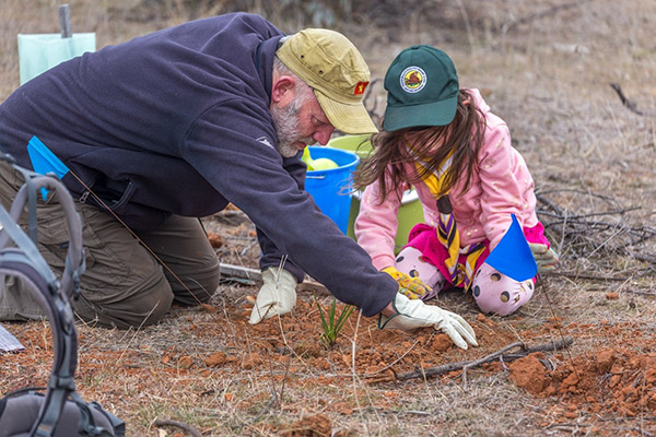 A male adult volunteer helps a female child prepare the ground for a plant to go in.
