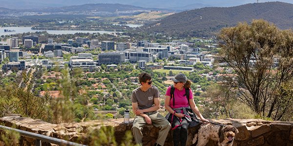 Two people sitting with their dog on a rock wall with a view across Canberra behind them.